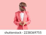 Small photo of Portrait of a funny bizarre man wearing a goofy rubber dinosaur mask and a trendy pink suit with a bowtie standing isolated on a pink colour background