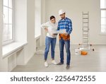 Small photo of Finishing painting decorating work foreman builder supervisor in hardhat showing young woman homeowner renovation plan standing in empty house or apartment with ladder and plaster bucket in background