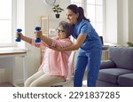 Small photo of Young nurse or physiotherapist in scrubs helping a happy retired old woman do fitness exercises with light weight dumbbells at home. Concept of physiotherapy for seniors
