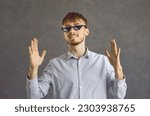 Small photo of Funny guy with ginger hair having fun. Studio headshot portrait of happy young man in cool pixelated thug life meme glasses standing against grey background