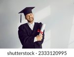 Small photo of Portrait of happy male graduate student standing with diploma. Positive young man wearing graduation robe and mortarboard smiling at camera standing at gray wall. Graduation, education concept