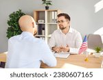 Small photo of Male Consul of United States of America sitting at office table with American flag, talking to young man about his immigration plans and listing what he needs to do for USA travel visa application
