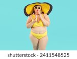 Portrait of fat woman in yellow swimsuit, beach hat and sunglasses drinking orange juice cocktail and looking cheerful at camera on blue background. Summer holiday trip and vacation concept.