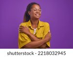 Young happy beautiful African American woman millennial with curly hair hugs herself to cheer up and get rid of loneliness dressed in casual yellow shirt, stands on isolated purple background.