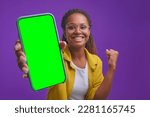 Small photo of Young overjoyed attractive African American woman gamer brag about completing game on mobile phone or with high scores on IQ test app stands in purple studio. Smartphone green screen
