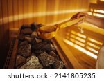 Woman in Finnish sauna, holding wooden sauna bucket. Woman is pouring water into hot stone in Sauna spa room. Woman in sauna - real, authentic moment.