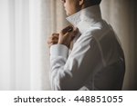 Man buttons up his white shirt standing in the front of a bright window