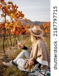 Small photo of Woman with poncho and hat sitting on blanket and drinks white wine. Relaxation in fall vineyard
