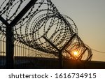 Small photo of Razor wire. Barbed tape on a fence at sunset. Rusty metal barbered wire on former border check point. Wire fence near old military barracks. Close up prison barber wire near jail or concentration camp