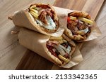 Small photo of Shawarma Gyro pita on wooden table. Greek food, sliced meat, potato, tomato and tzatziki, paper wrap, close up view.