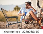Handsome man with sunglasses and drink sitting in camper chair in front of camper rv, looking into distance and smiling. Camping, outdoors, roadtrip, nature concept.