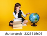 Small photo of Cute tittle schoolgirl pointing to world globe while sitting near bunch of books, isolated over yellow background, copy space. Back to school concept