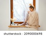 Home comfort.Lifestyle portrait of young blond woman with bunch hairstyle sitting on windowsill in lightroom. Cute bunch hairstyle lady enjoy winter holiday sitting, holding cup of coffee.