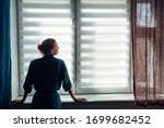 Small photo of Woman's silhouette at window in the background light. Young woman in gown stands near window with blinds. Quarantine, self-isolation, stay home, self-preservation, coronavirus pandemic.