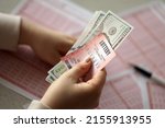 Small photo of A young woman holds the lottery ticket with complete row of numbers and dollar bills on the lottery blank sheets background. Lottery winner concept