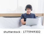Happy asian businessman working from home. Handsome young man sitting and wear headphone with talking by video conference on bed. Male wearing earphone and enjoy listening music by laptop computer.