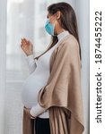 Small photo of Young pregnant woman is standing by window with face protective mask. She is worried about childbirth because od Covid-19 pandemic.