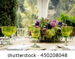 Formal table setting for a wedding with floral centerpiece and colorful green glasses in front of a large view window overlooking a garden