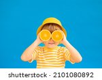 Small photo of Happy child holding slices of orange fruit like sunglasses. Kid wearing striped yellow t-shirt against blue paper background. Healthy eating and summer vacation concept