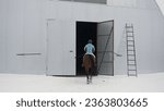 Small photo of Hangar for racing in winter. Girl rider on a black horse in winter. Indoor hangar for training horses. A young woman rides a horse in a covered hangar.