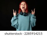 Cool young redhead girl in a blue oversized hoodie shouting with crazy expression doing rock symbol with hands up. Music star. Heavy concept. Sign of the horns, tongue is out. isolated on black.