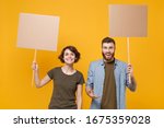 Small photo of Smiling protesting young two people guy girl hold protest signs broadsheet blank placard on stick isolated on yellow background studio portrait. Protests strikes pickets concept. Youth against city