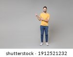 Full length young caucasian smiling cheerful bearded attractive happy man in casual yellow basic t-shirt point index finger aside on copy space area isolated on grey color background studio portrait