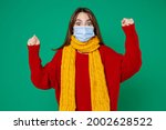 Excited young woman in red sweater yellow scarf sterile face mask to safe from coronavirus virus covid-19 during pandemic quarantine doing winner gesture isolated on green background studio portrait