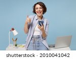 Young happy fun secretary employee business woman in casual shirt work stand at white office desk with pc laptop hold mobile cell phone show thumb up gesture isolated on pastel blue background studio.