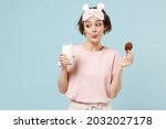 Young excited fun woman 20s wear pajamas jam sleep eye mask rest relaxing at home hold glass of milk with cookie isolated on pastel blue background studio. Breakfast lifestyle night bedtime concept