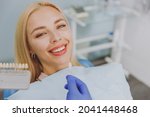 Small photo of Close up young smiling woman choosing veneers enamel color palette with stomatologist compare teeth shade sit at dentist office chair indoor light modern cabinet Healthcare enamel whitening treatment