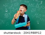 Young male kid school boy 5-6 years old in t-shirt backpack biting sandwich eat lunch at break isolated on green wall chalk blackboard background. Childhood children kids education lifestyle concept.
