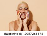 Small photo of Young smiling happy fun blond latin gay man 20s wearing beige tank shirt posing with make up fingers painted in rainbow flag colors hold face isolated on plain light ocher background studio portrait.