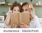 Small photo of Chef cook baker mom woman in white shirt work baby girl helper read cookbook point finger up hide at kitchen table home Cooking food process concept Mommy little kid daughter prepare fruit sweet cake