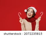 Young smiling fun happy woman 20s wear Santa Claus Christmas red hat hold candy cane stick lollipop look aside isolated on plain red background studio portrait. Happy New Year 2022 celebration concept