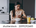 Young confused shcoked sad housewife woman 20s in t-shirt eat breakfast in morning look at smart watch check time hurrying rush cooking food in light kitchen at home Healthy diet lifestyle concept.