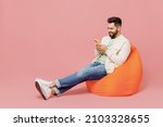 Full body young smiling cheerful happy man 20s in trendy jacket shirt sit in bag chair hold in hand use mobile cell phone isolated on plain pastel light pink background studio People lifestyle concept