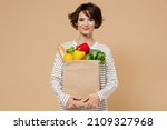 Small photo of Young smiling caucasian cheerful happy fun vegetarian woman 20s in casual clothes hold paper bag with vegetables after shopping look camera isolated on plain pastel beige background studio portrait