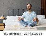 Small photo of Young peaceful man in casual blue t-shirt sitting on bed in lotus pose yoga om aum gesture relax meditate try to calm down listen music in earphones rest relax spend time in bedroom home in own room