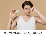 Small photo of Attractive sad young man 20s perfect skin in undershirt hold contact lenses case point finger on container rub his eyes isolated on plain pastel beige background. Eyecare healthcare procedures concept