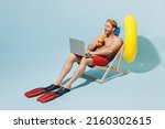 Full length young man 20s in red shorts swimsuit sit in deckchair inflatable ring hold cocktail use laprop pc computer work isolated on pastel blue background Summer vacation sea rest sun tan concept.