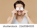 Small photo of Attractive joyful young man 20s perfect skin in undershirt hold cover eyes with cotton pad isolated on light pastel beige background studio portrait. Skin care healthcare cosmetic procedures concept.