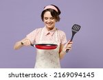 Young smiling happy cheerful housewife housekeeper chef cook baker woman in pink apron holding red frying pan spatula isolated on pastel violet background studio portrait Cooking food process concept.