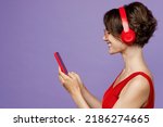 Side view young smiling woman 20s she wear red tank shirt eyeglasses headphones listen to music hold in hand use mobile cell phone isolated on plain purple background studio. People lifestyle concept