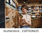 Small photo of Young satisfied woman 20s in casual clothes shopping at supermaket grocery store buy choosing white wine alcohol hold bottle prop up chin inside hypermarket. People purchasing gastronomy food concept