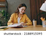 Young smiling happy cheerful fun european successful employee business woman 20s she wearing casual yellow shirt writing in notebook diary sit work at wooden office desk. Achievement career concept