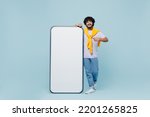 Small photo of Full size young bearded Indian man 20s wears white t-shirt stand near pointing on big mobile cell phone with blank screen workspace area isolated on plain pastel light blue background studio portrait