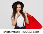 Small photo of Young woman wear t-shirt hat hold in hand red paper package bags after shopping say hush shhh be quiet finger on lips isolated on plain solid white color background. Black Friday sale buy day concept