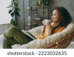 Sideways young woman wear casual clothes drink coffee read novel book sits in armchair stay at home flat rest relax spend free spare time in living room indoor grey wall