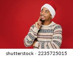 Merry young man wear warm Christmas sweater Santa hat posingl look aside on workspace area mock up prop up chin isolated on plain red color background. Happy New Year 2023 celebration holiday concept